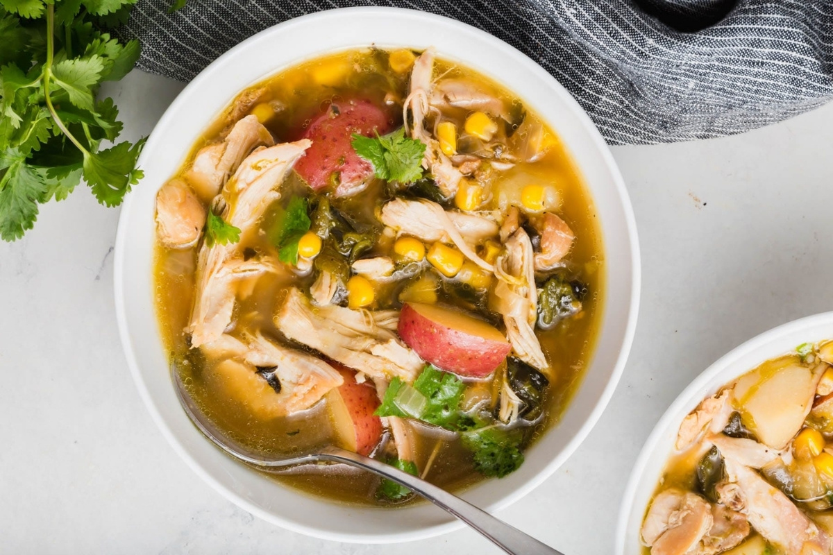 A bowl of poblano chicken soup with shredded chicken, corn, red potatoes, spinach, and fresh cilantro, served with a spoon on a white marble surface next to a gray cloth napkin and fresh cilantro.