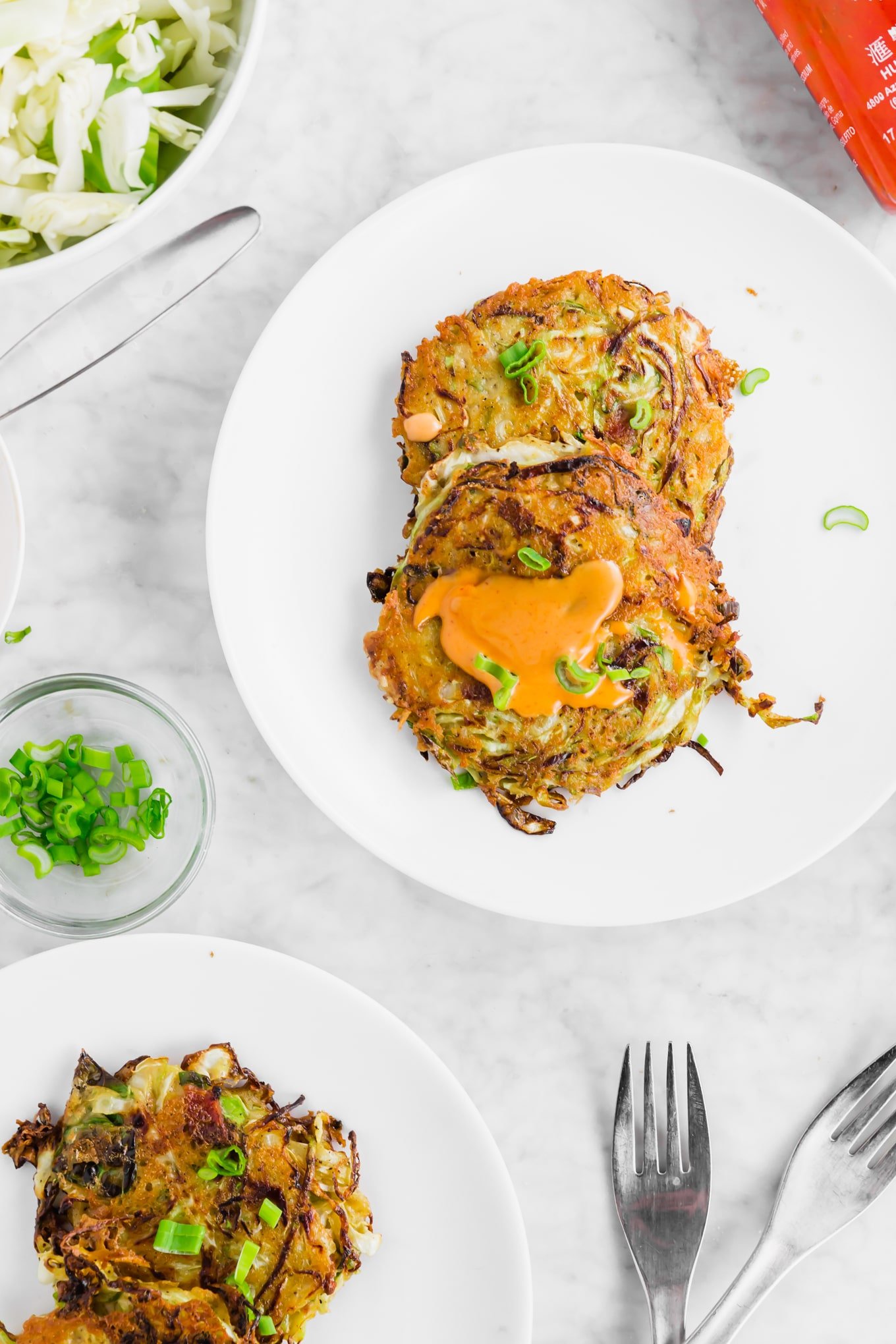 A photo of cabbage bacon fritters on a plate topped with spicy sriracha mayo and green onions. 