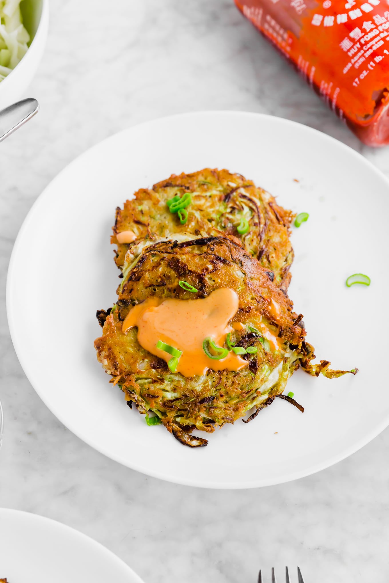 A photo of cabbage bacon pancakes on a plate topped with green onions and sriracha mayo. 