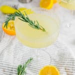 A coupe glass filled with meyer lemon rosemary gin cocktail with a fresh sprig of rosemary and sliced meyer lemons around the glass.