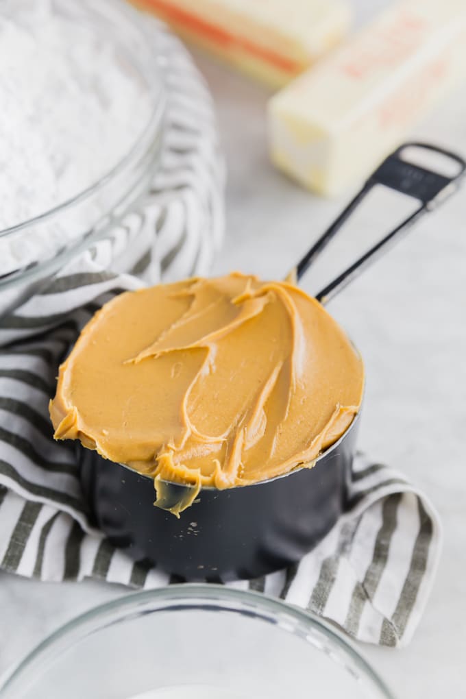 A measuring cup filled with peanut butter with a bowl of powdered sugar and sticks of butter to make peanut butter frosting. 