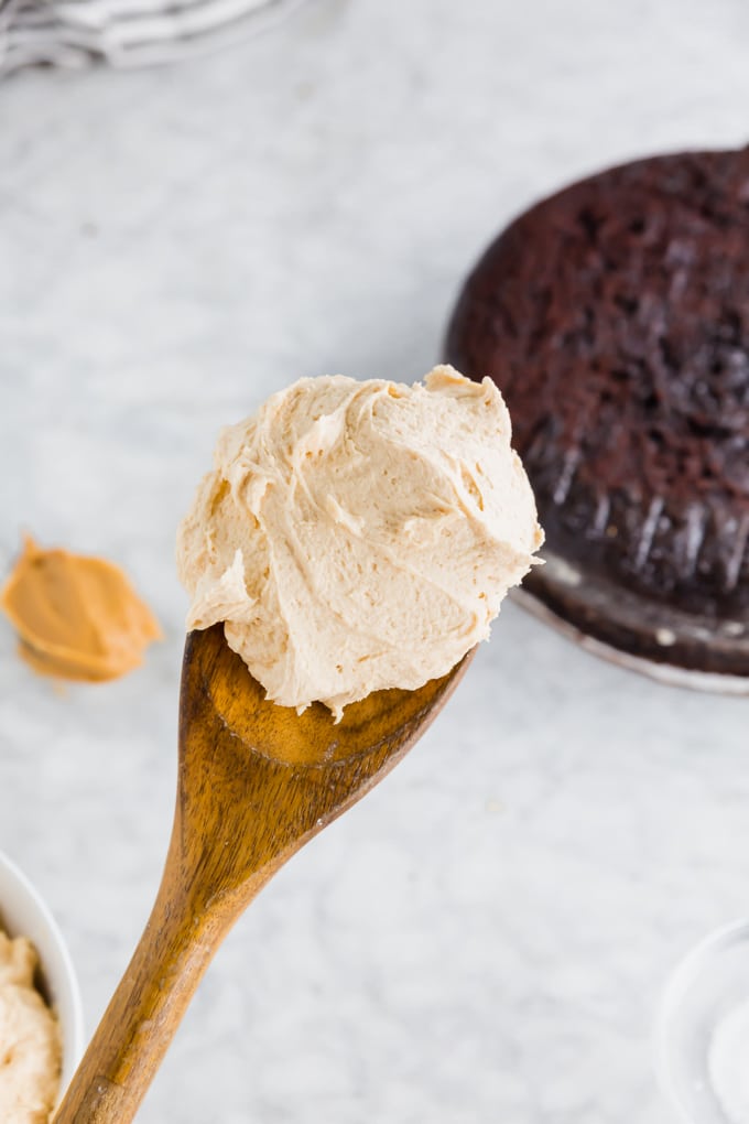 A photo of a wood spoon with peanut butter frosting on it with a gluten-free chocolate cake in the background. 