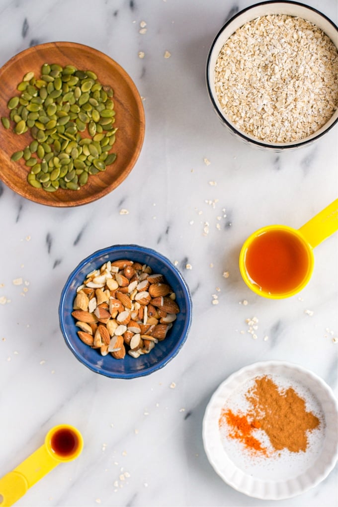 An overhead view of bowls of pumpkin seeds, oats, almonds, maple syrup, cayenne, and vanilla extract.