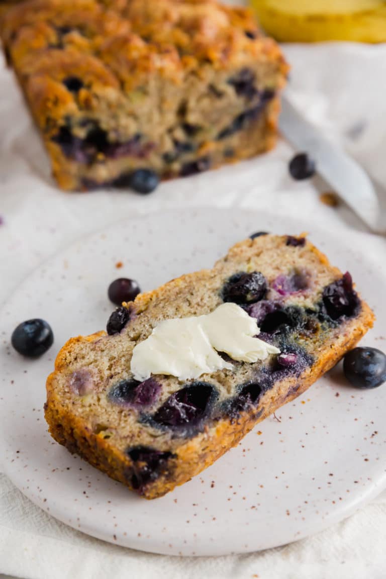 A photo of a slice of gluten-free vegan blueberry banana bread with vegan butter on top with a loaf of bread in the background.