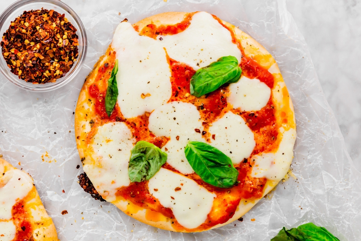 A mini Margherita pita pizza topped with melted mozzarella, tomato sauce, and fresh basil leaves, sits on crumpled parchment paper with a small bowl of red pepper flakes nearby.