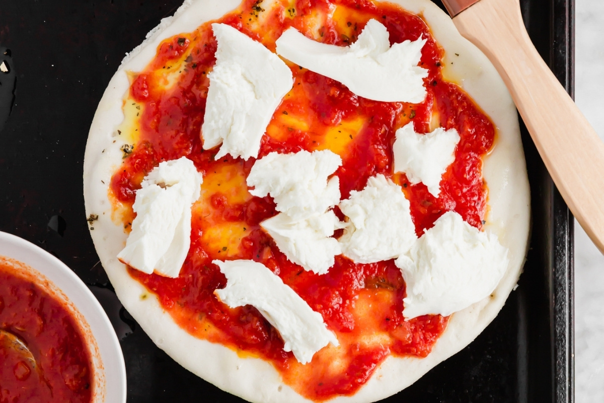 A round pita pizza dough topped with tomato sauce and uneven pieces of fresh mozzarella cheese, ready to be baked, sits on a black tray next to a bowl of extra tomato sauce.