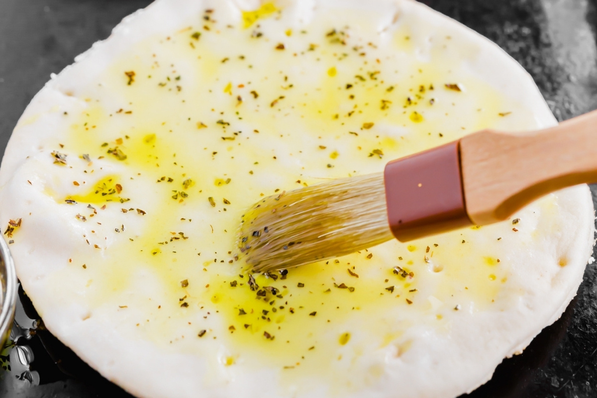 A pastry brush spreads olive oil and herbs onto a round, uncooked pita pizza dough on a dark surface.