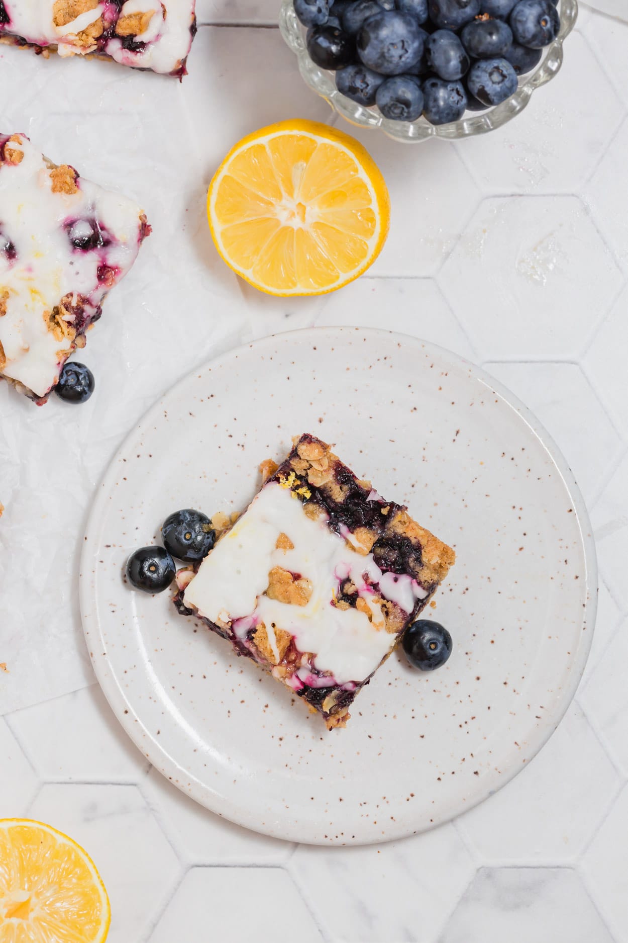 An overhead photo of gluten-free vegan blueberry crumble bar on a plate with fresh blueberries and lemon halves surrounding it.