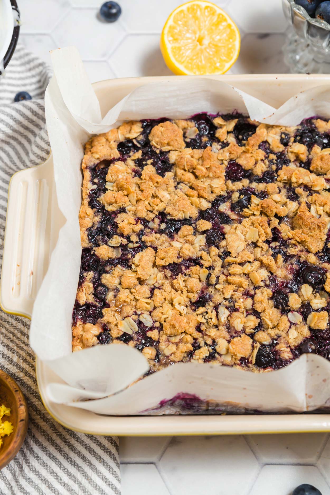 A photo of gluten-free vegan blueberry crumb bars baked in an 8x8-inch baking dish straight from the oven with fresh blueberries and lemon surrounding it. 