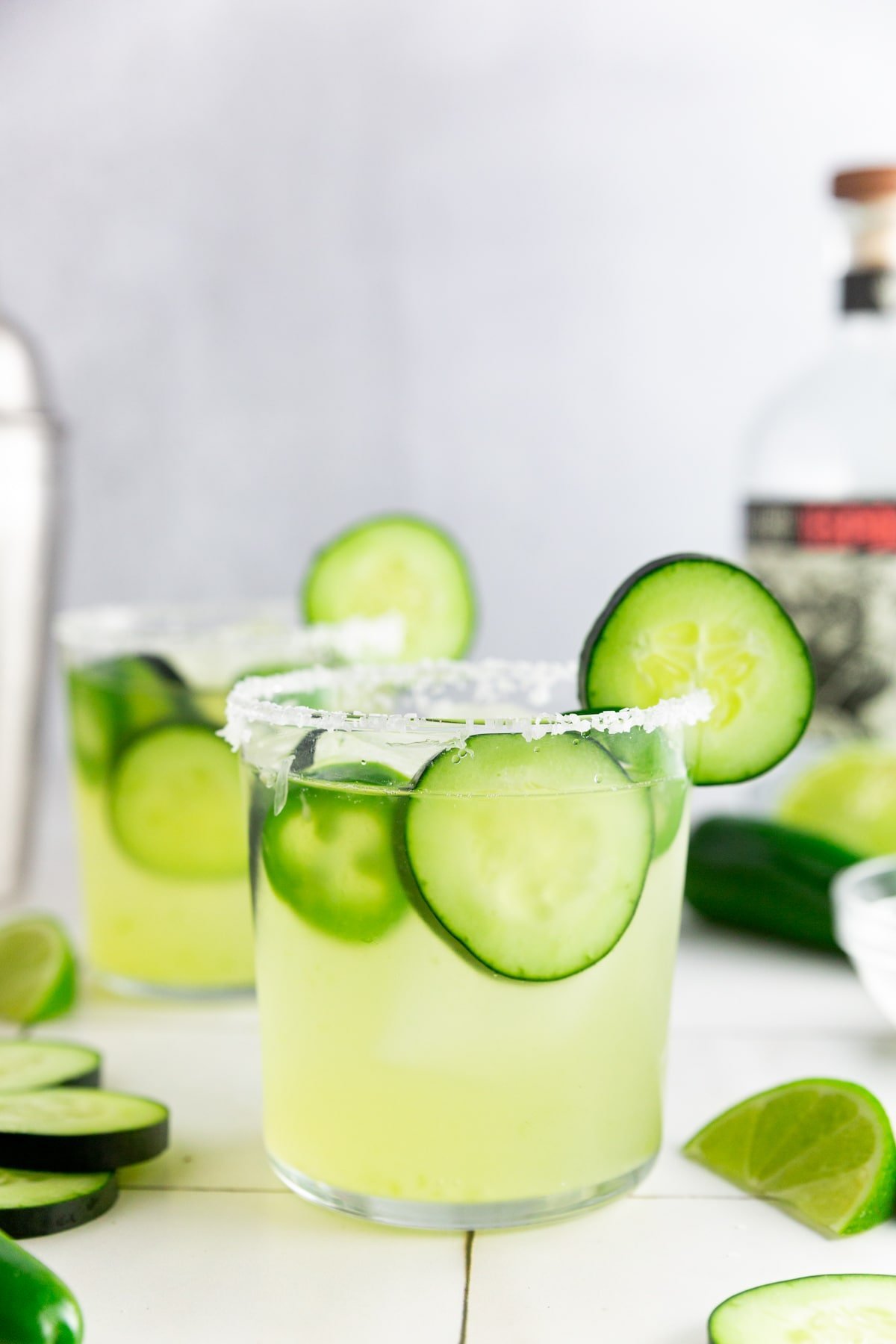 A photo of a cucumber jalapeño margarita with a salted rim and a bottle of tequila and a cocktail shaker in the background.