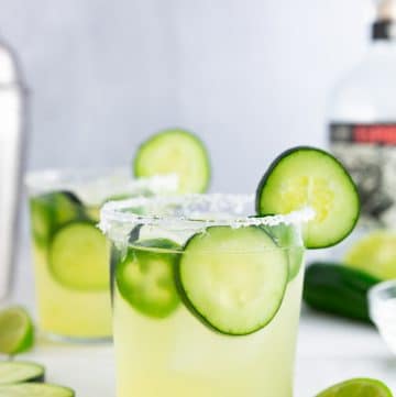 A photo of a cucumber jalapeño margarita with a salted rim and a bottle of tequila and a cocktail shaker in the background.