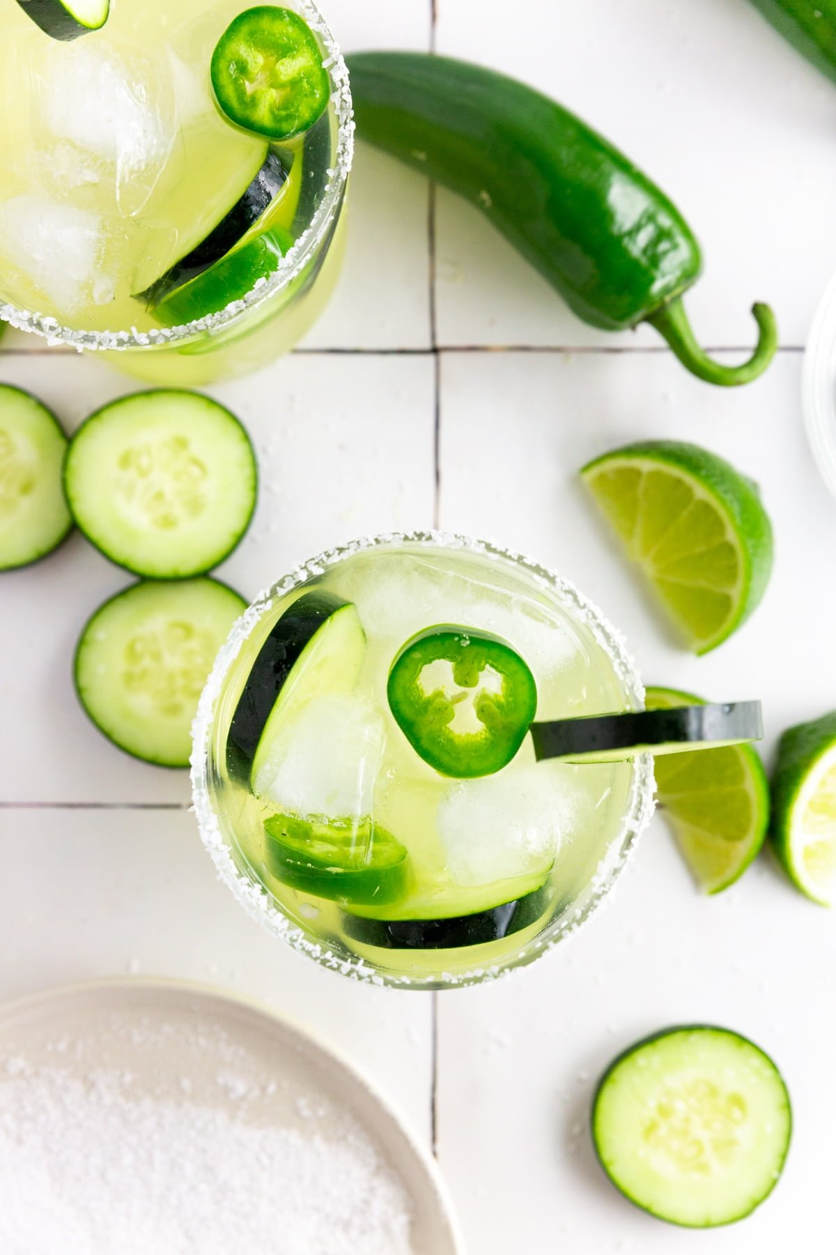 An overhead photo of two margarita glasses filled with jalapeño cucumber margaritas with sliced jalapeño and cucumbers in it and lime wedges on a table.