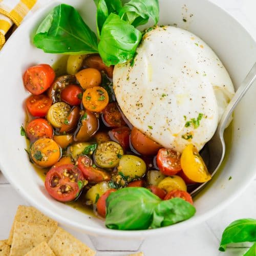 An overhead photo of a serving bowl with marinated cherry tomatoes, burrata and fresh basil with a serving spoon.