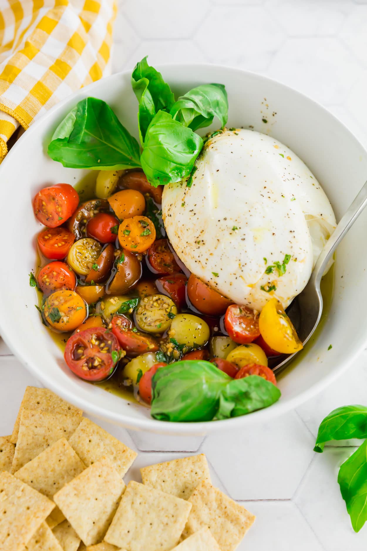 An overhead photo of a serving bowl with marinated cherry tomatoes, burrata and fresh basil with a serving spoon.