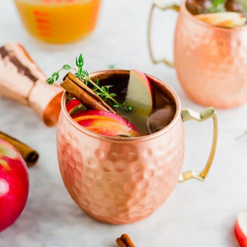 A copper mug filled with apple cider thyme moscow mule with a cinamon stick and apples on top.