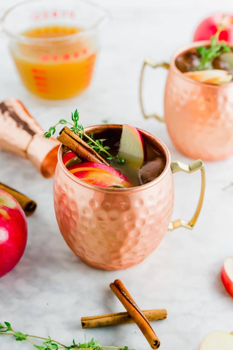 A copper mug filled with apple cider thyme moscow mule with a cinamon stick and apples on top.