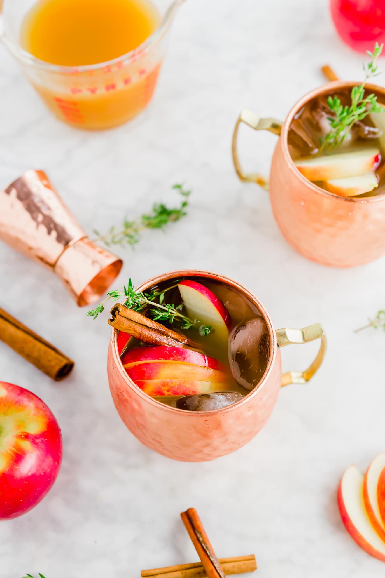 two copper mugs on a marble table filled with apple cider cocktail with a rose gold cocktail jigger and pink lady apples surrounding it.