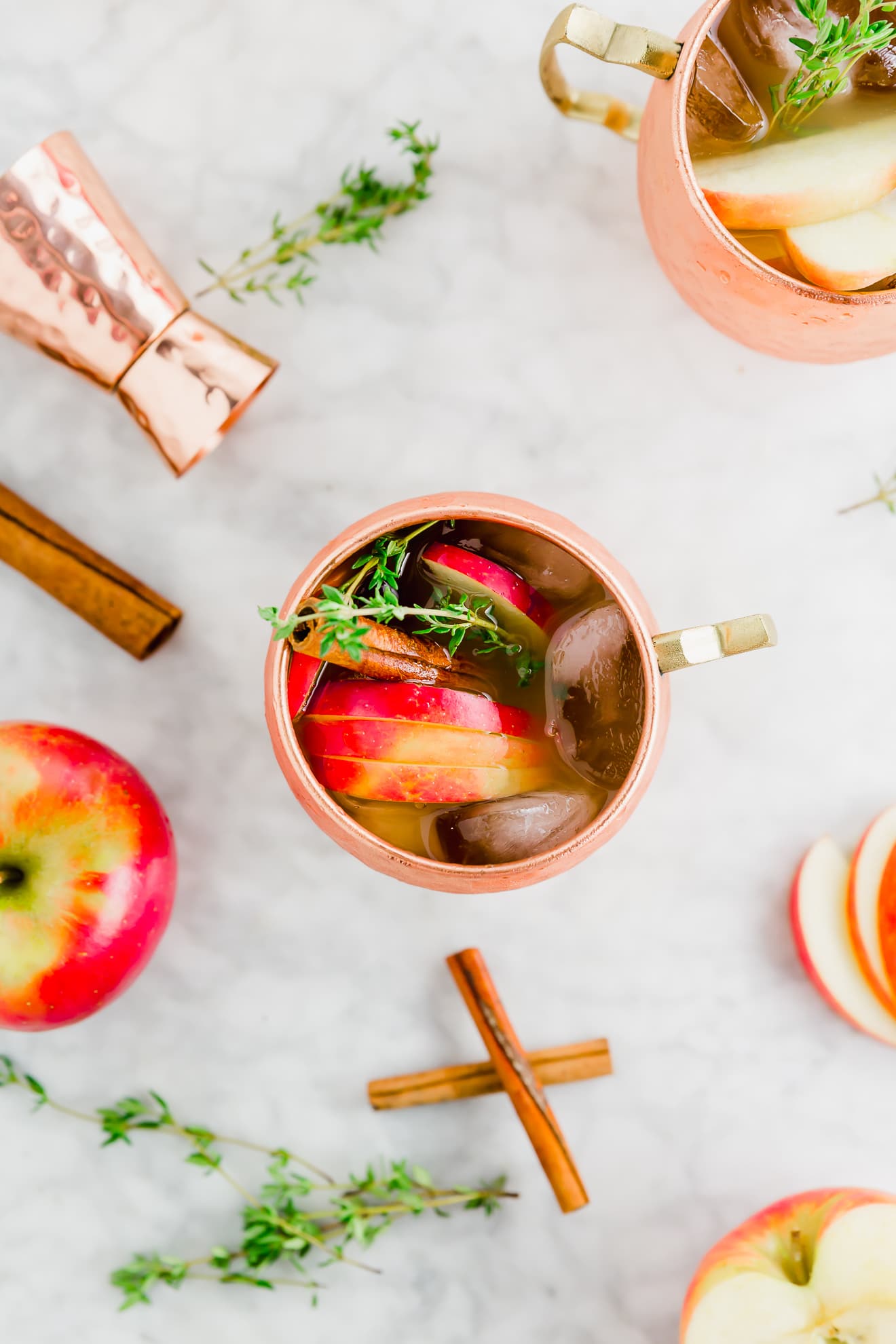 an aerial photo of two glasses of apple cider moscow mule topped with fresh thyme, sliced honeycrisp apples and cinnamon sticks.