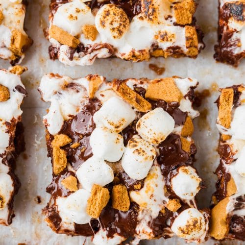 An overhead view of sliced gluten-free smores bars topped with graham crackers and marshmallows on a piece of parchment paper.