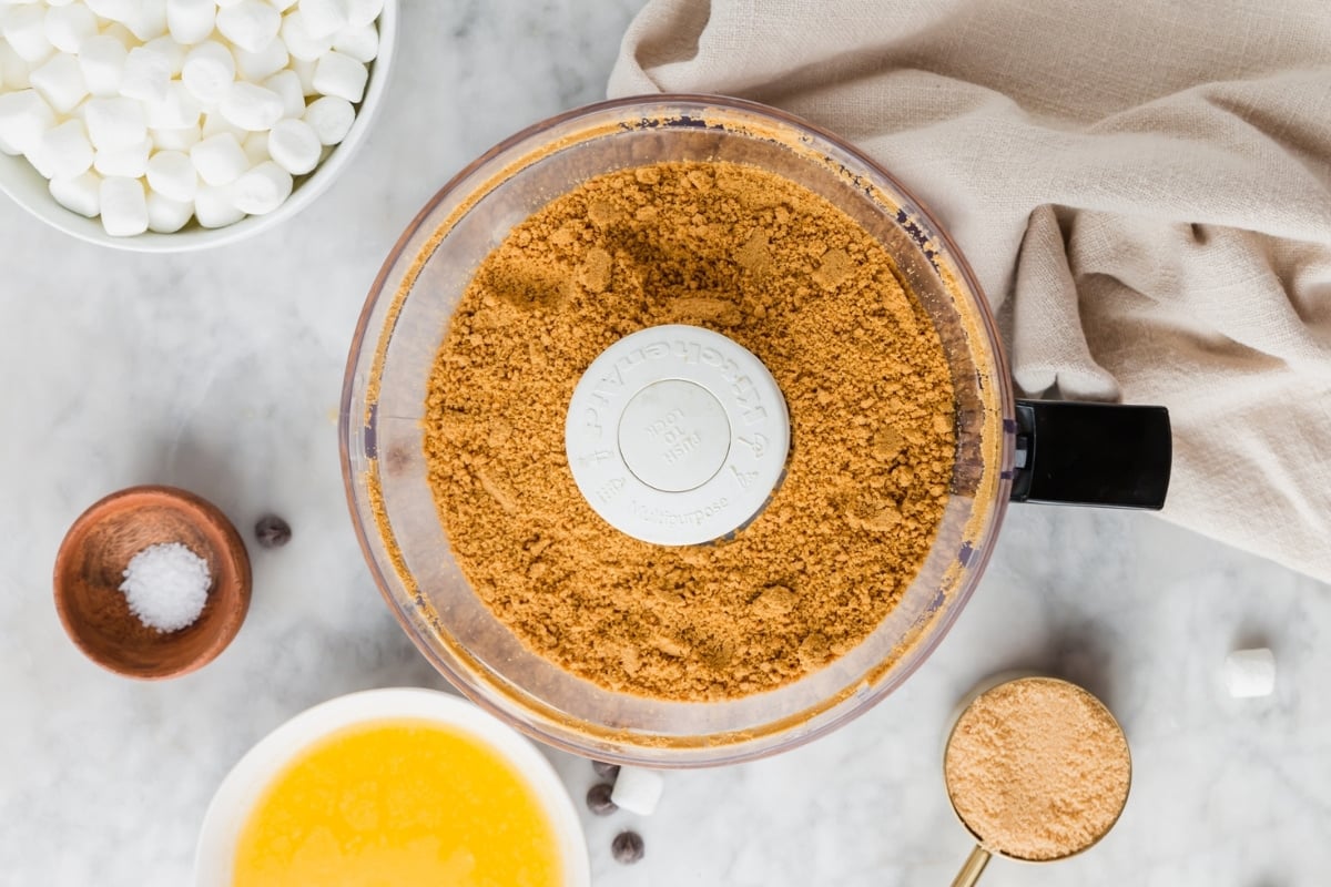 Food processor with crushed graham crackers for Gluten-Free S’mores Bars, surrounded by melted butter, marshmallows, salt, a measuring cup of crumbs, and a beige cloth on a light countertop.