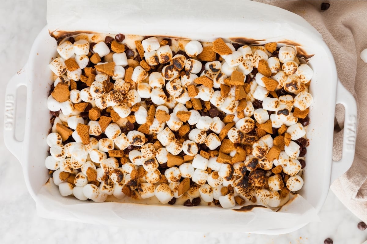 A white baking dish filled with toasted mini marshmallows, graham cracker pieces, and melted chocolate to create delicious Gluten-Free S’mores Bars on a marble surface.