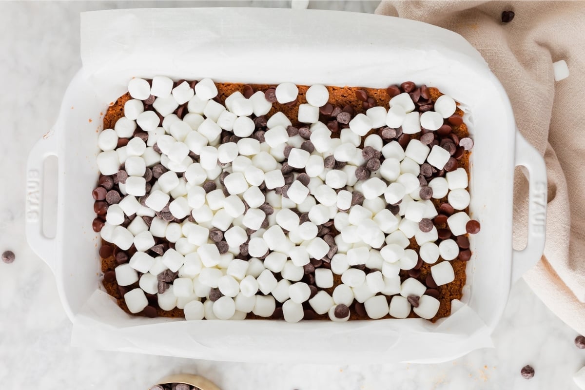 A white baking dish lined with parchment paper, filled with Gluten-Free S’mores Bars topped with mini marshmallows and chocolate chips, sits on a light marble surface.