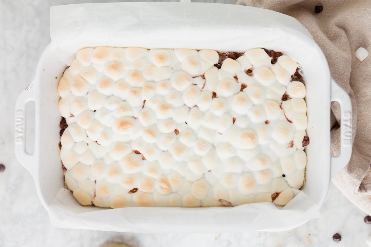 A white baking dish filled with Gluten-Free S’mores Bars, topped with golden-brown toasted mini marshmallows, sits on a light surface.