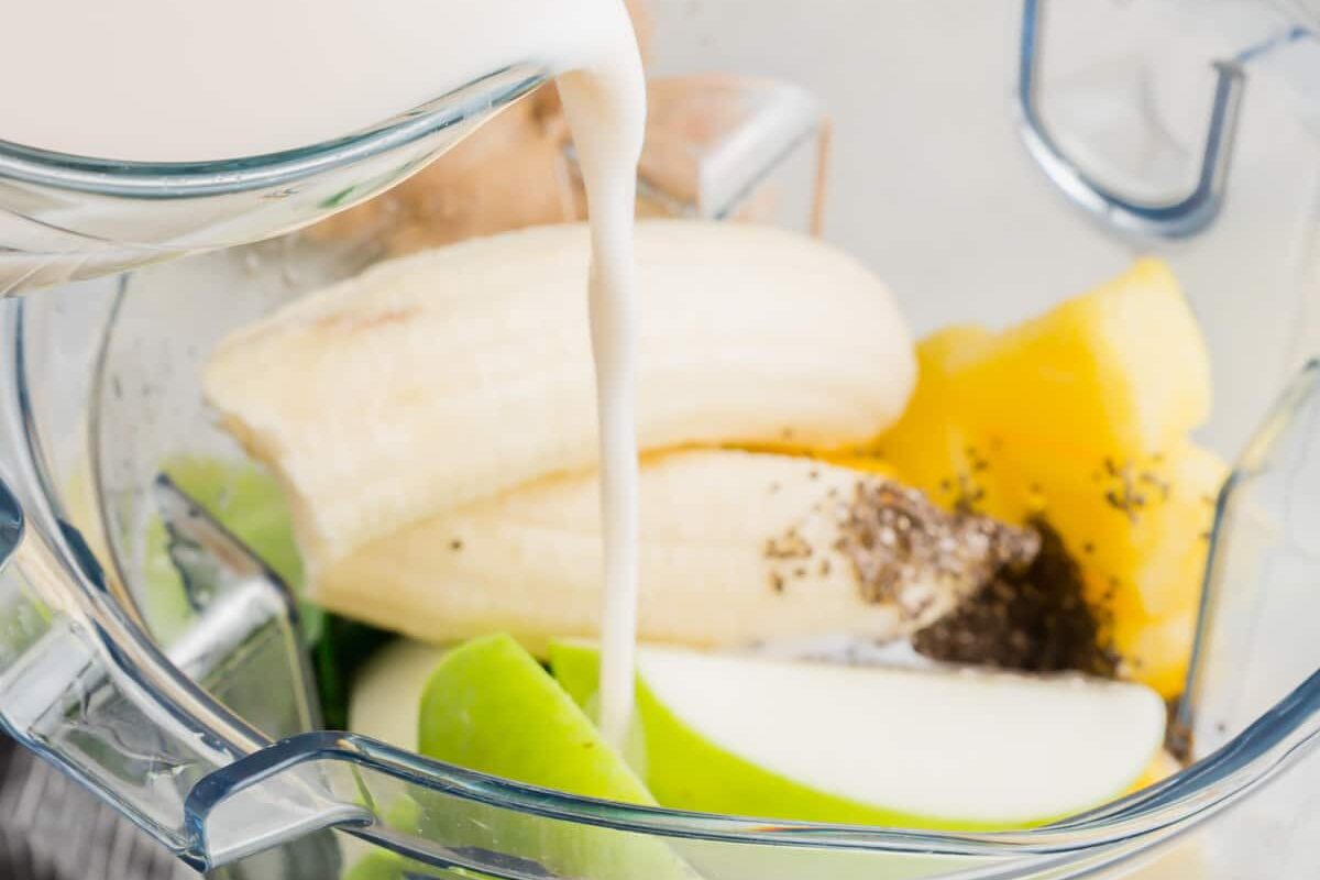 Almond milk being poured into a blend with apple, banana, pineapple and kale.