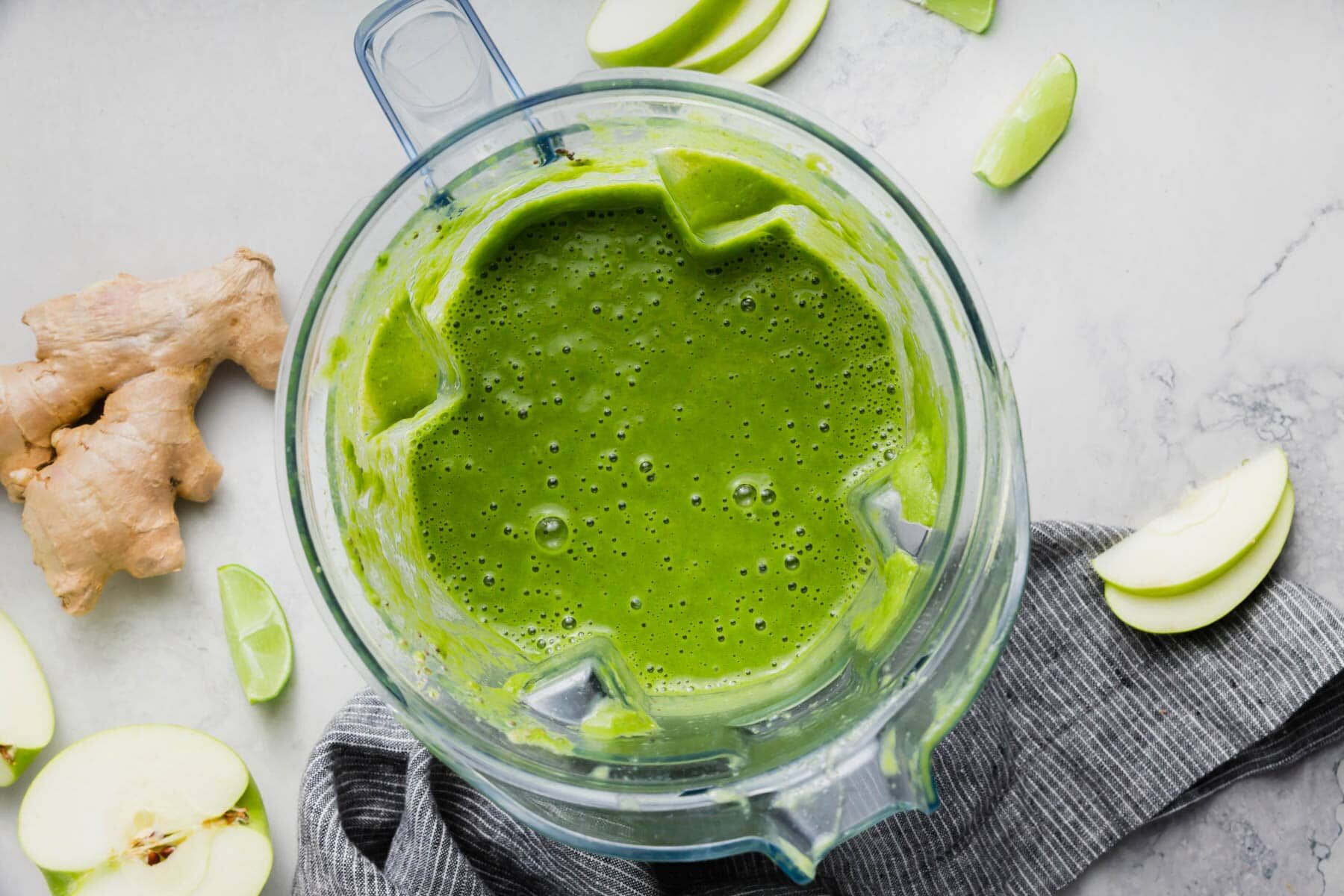 A blender with apple kale green smoothie in it and fresh ginger and apple on the table.