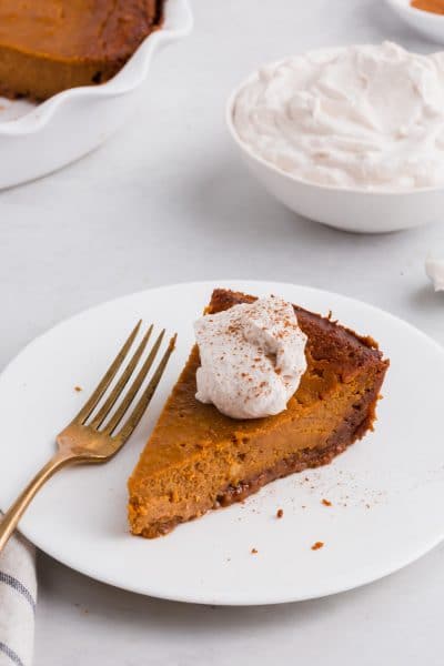 A photo of a piece of gluten-free pumpkin pie on a plate with a fork and a bowl of whipped cream in the background.