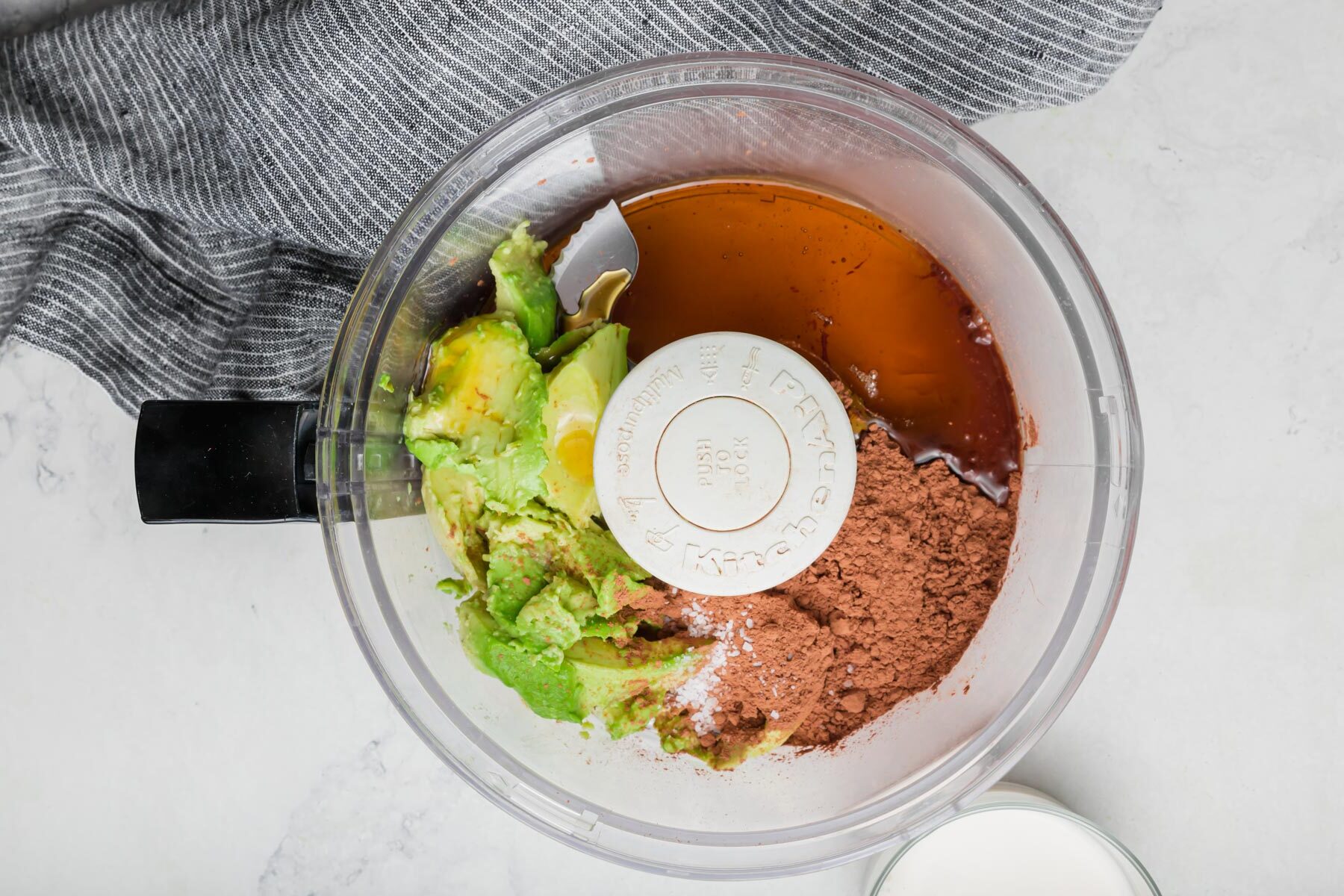 A food processor with maple syrup, cocoa powder, avocado, and salt in it.