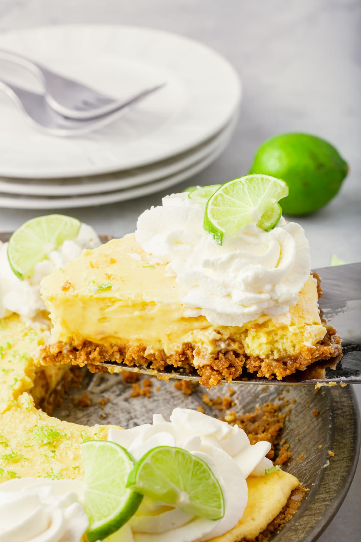 A piece of key lime pie with graham cracker crust being lifted out of a pie tin with a spatula.