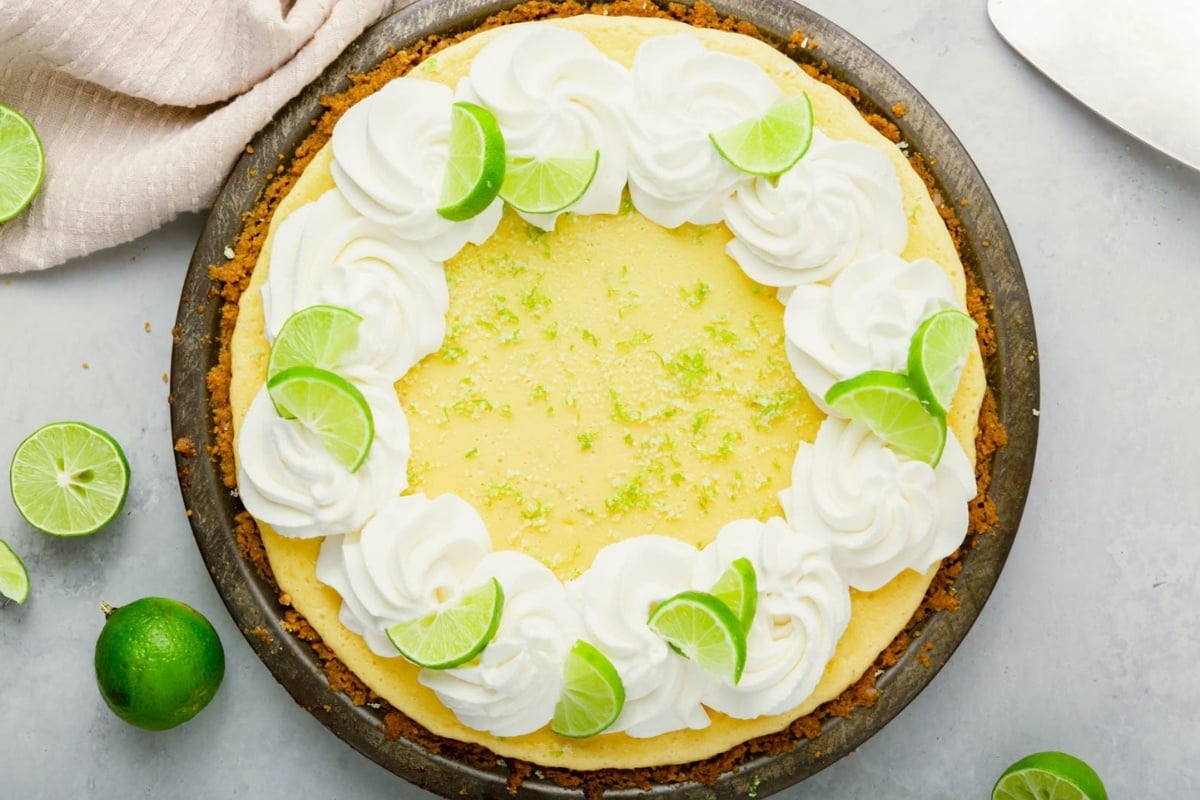 A gluten-free key lime pie with a graham cracker crust, topped with swirls of whipped cream and small lime wedges. Whole and sliced limes are scattered on the light gray surface nearby.