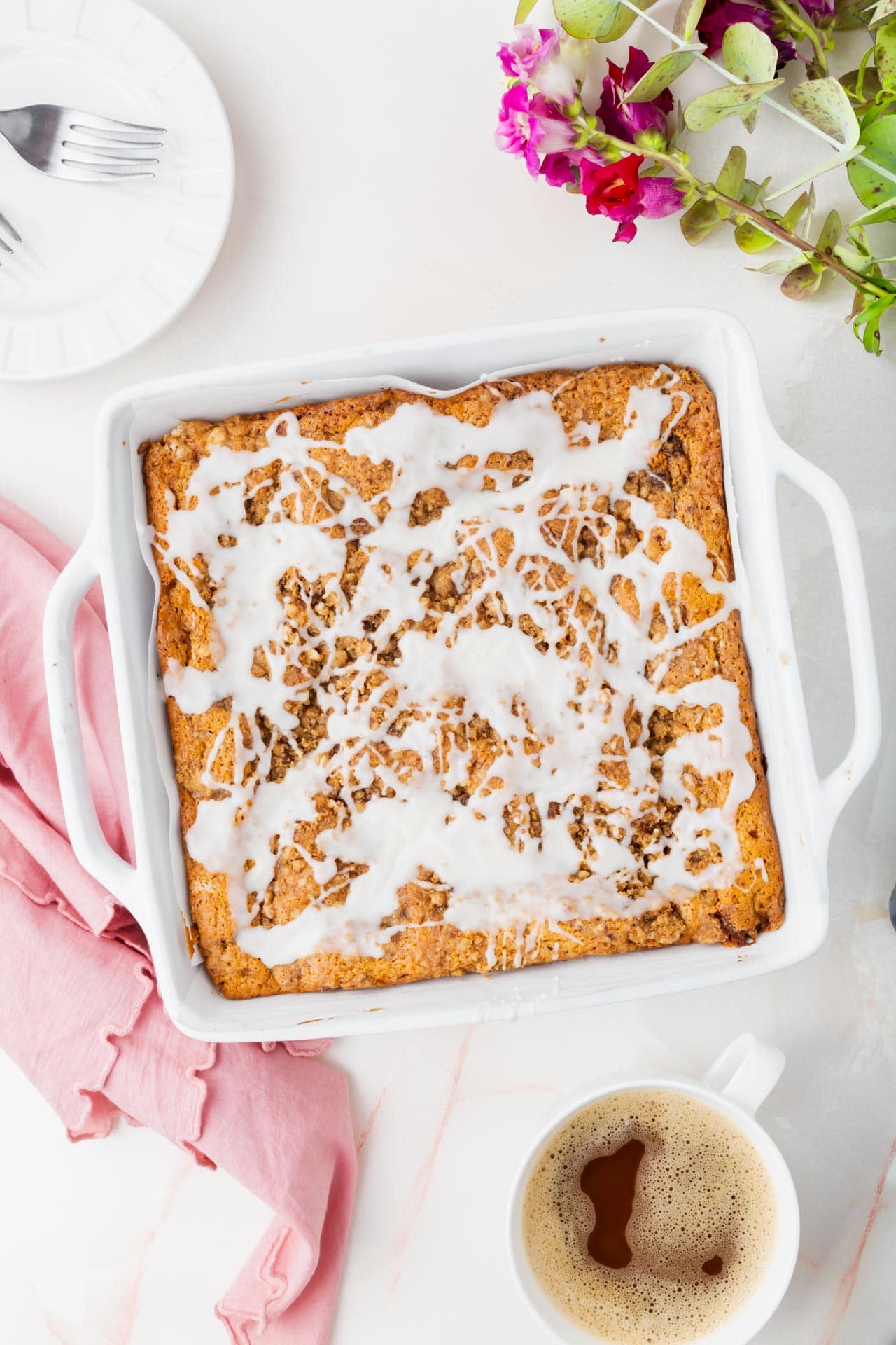 Overhead photo of coffee cake with glaze in a square baking dish with a cup of coffee and flowers.