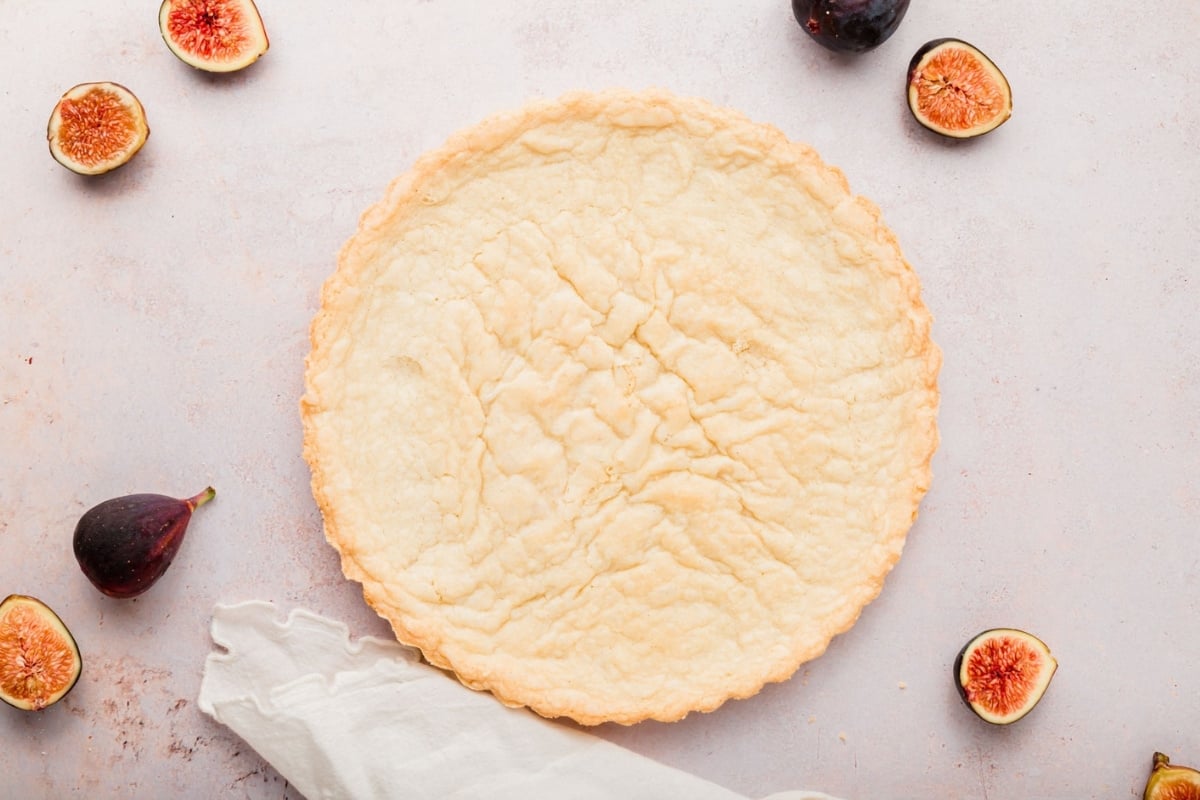 A gluten-free fig tart crust sits on a light surface surrounded by fresh figs, some whole and some halved. A white cloth is partially draped near the pie crust.