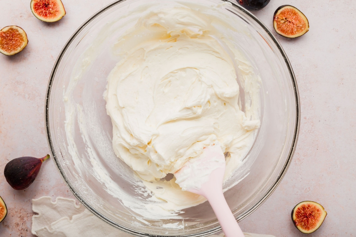 A glass bowl with a creamy whipped mixture and pink spatula rests beside halved figs on a light surface, perfect for preparing a delicious gluten-free fig tart.