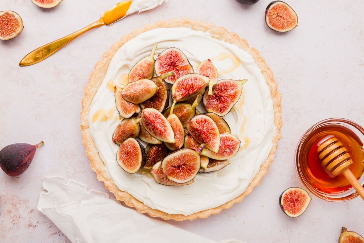 A gluten-free fig tart topped with sliced fresh figs on cream, surrounded by a honey dipper, bowl of honey, extra fig pieces, and a gold knife with cream on it, set on a light tabletop.