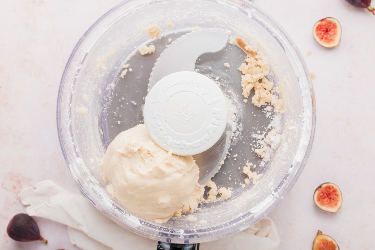 A food processor with gluten-free fig tart dough inside, surrounded by scattered figs on a light surface. Some dough and flour residue remains in the processor.