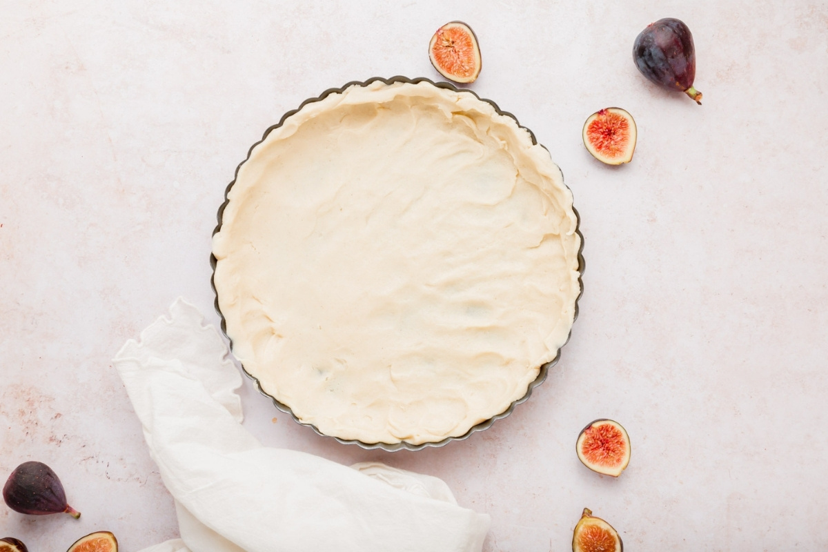 A tart pan filled with unbaked gluten-free fig tart crust sits on a light surface, surrounded by fresh figs, fig halves, and a white cloth.