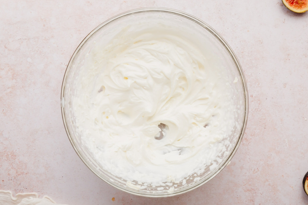 A glass bowl filled with freshly whipped cream sits on a light pink surface, ready to top a gluten-free fig tart.