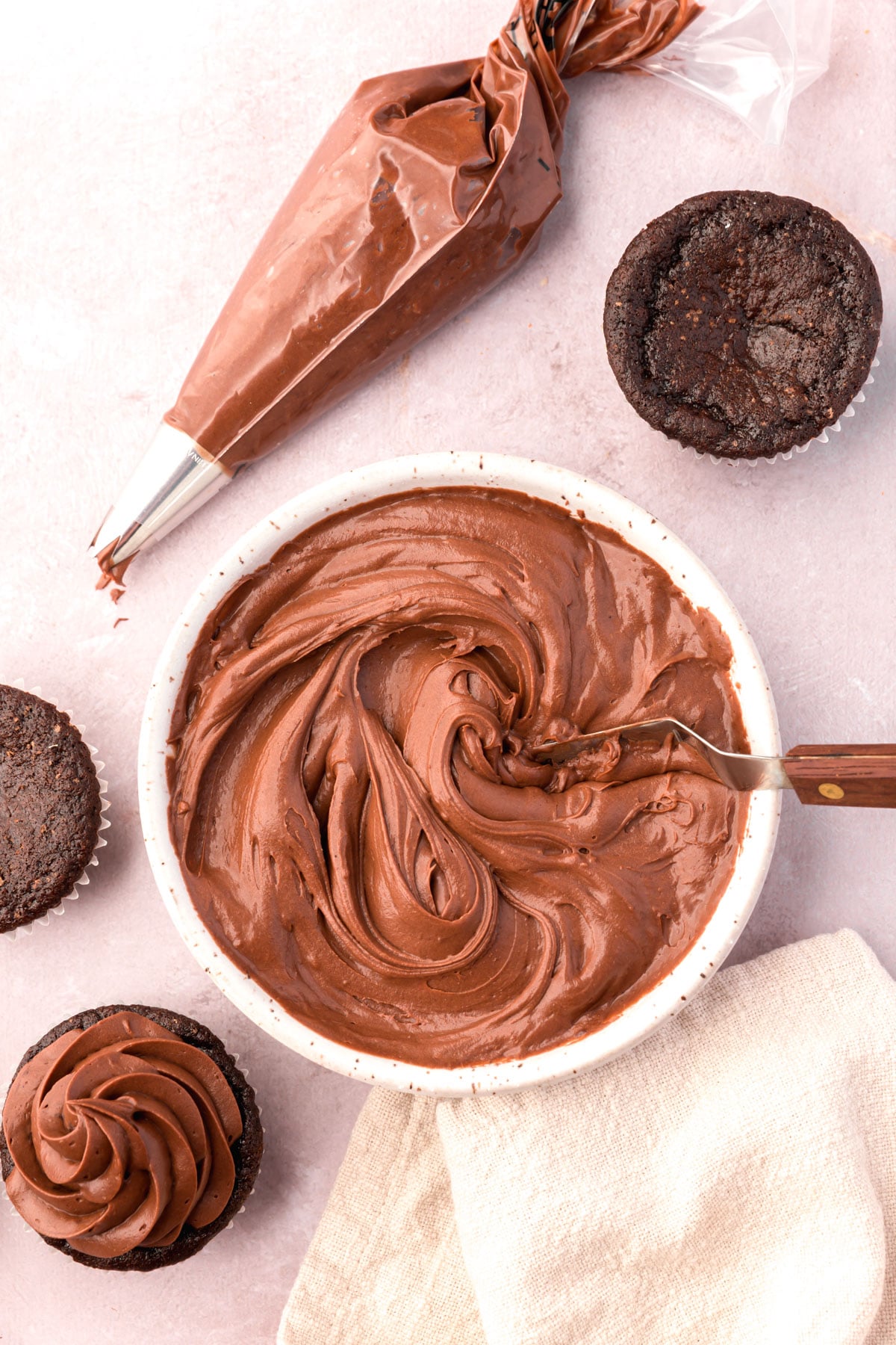 A bowl of chocolate cream cheese frosting surrounded by cupcakes and a piping bag.