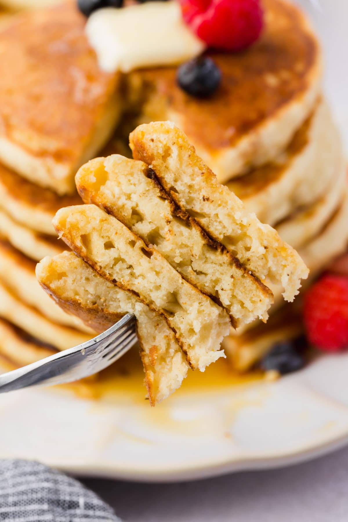 A forkful of gluten-free pancakes in front of a plate of a stack of gluten-free pancakes topped with butter, fresh berries, and maple syrup.