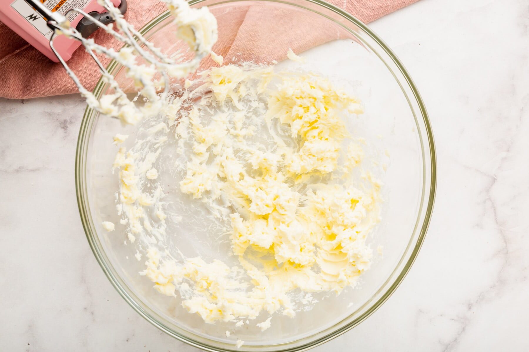 A glass mixing bowl containing creamed butter and sugar, with a hand mixer and a pink kitchen towel nearby on a marble countertop.