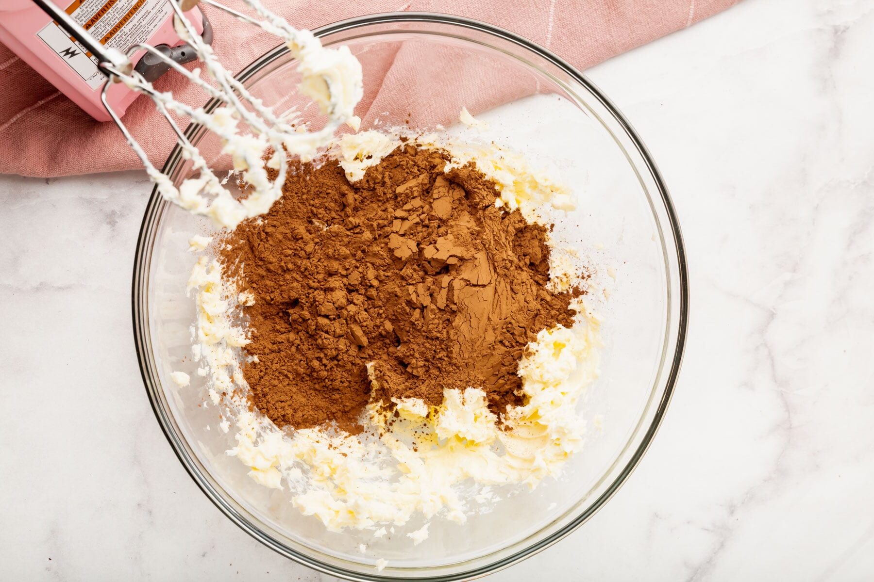 A glass mixing bowl with cocoa powder, sugar, and butter being mixed by an electric hand mixer on a white countertop next to a pink kitchen towel.