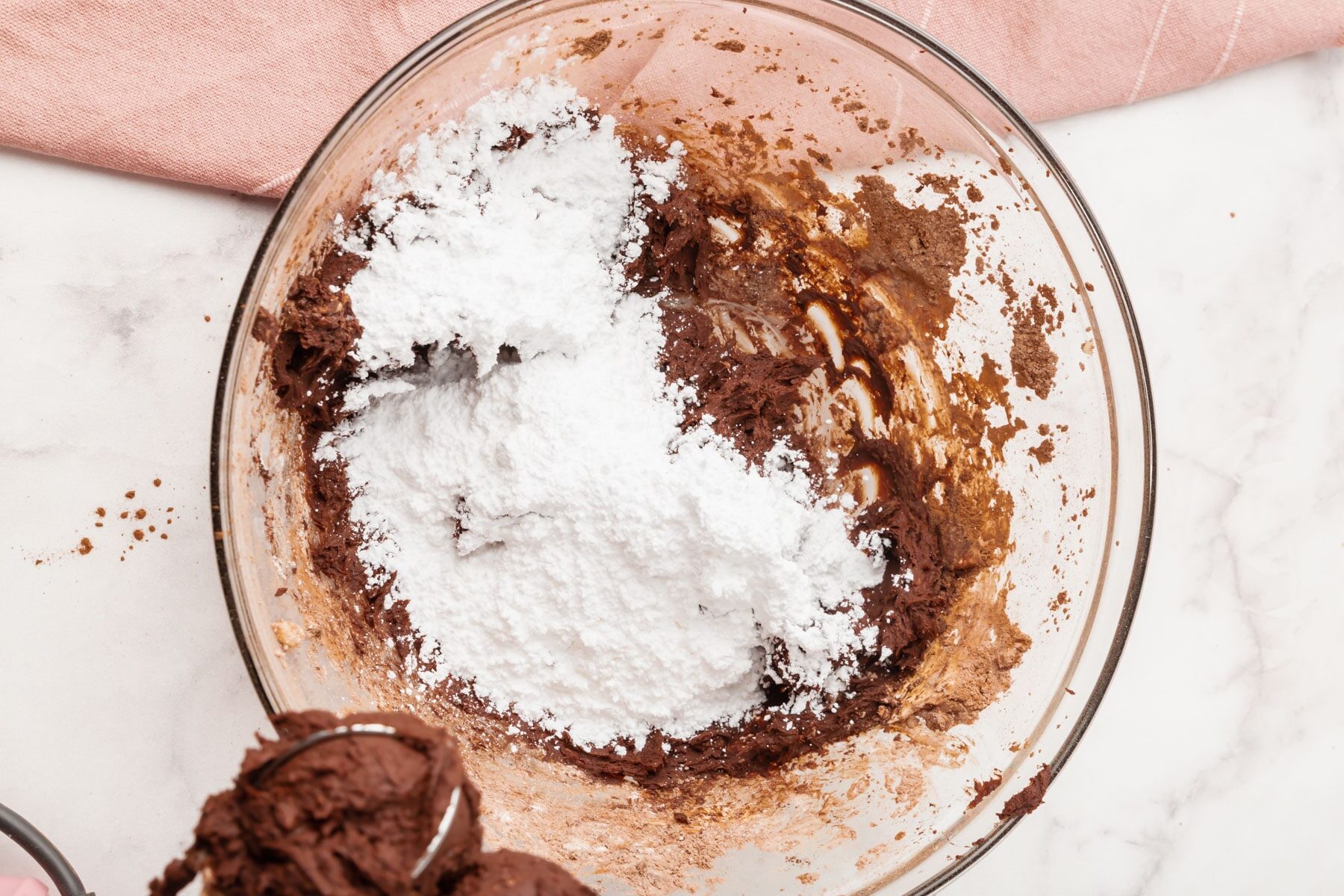 A glass bowl containing chocolate batter and a mound of powdered sugar on top, sitting on a marble surface with part of a pink cloth visible to the side.