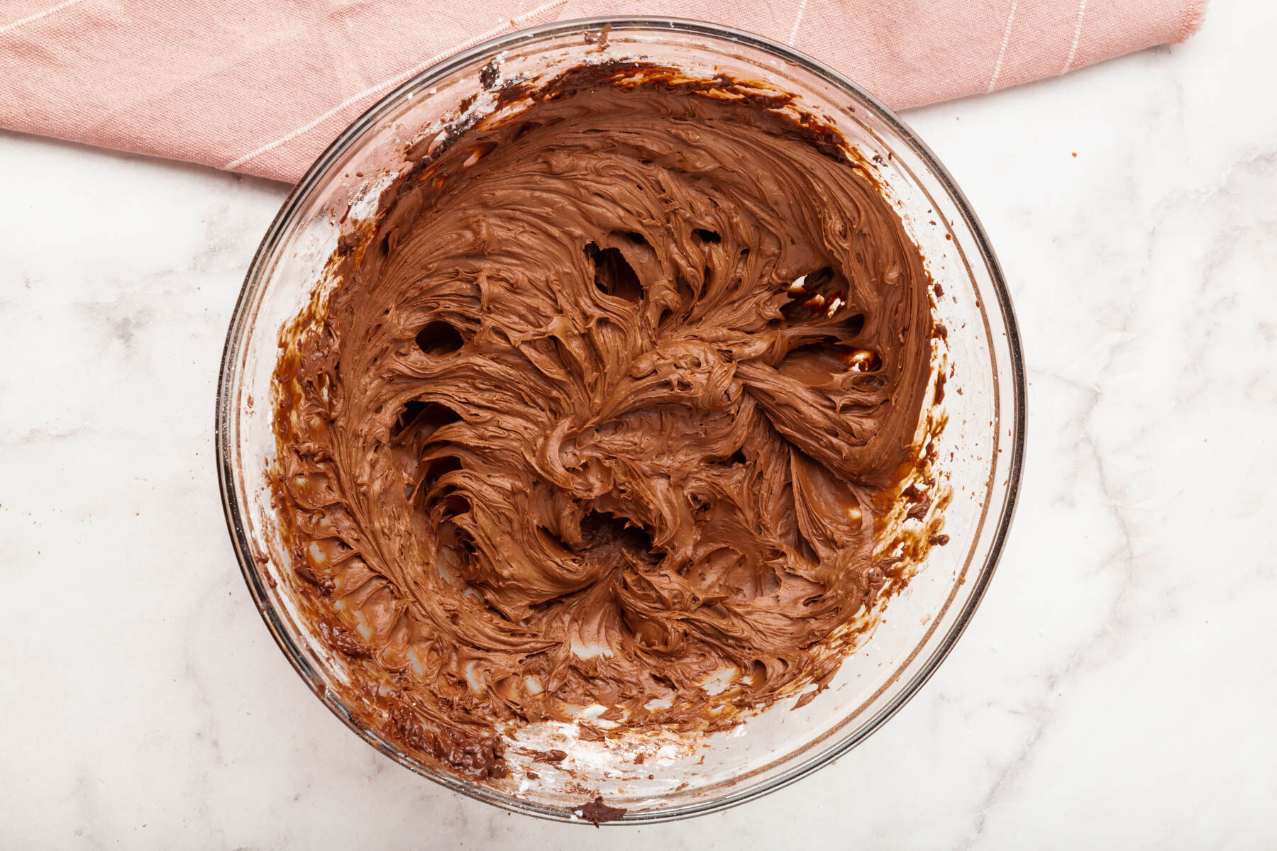 A glass bowl filled with creamy, swirled chocolate batter sits on a white marble surface next to a folded light pink cloth.