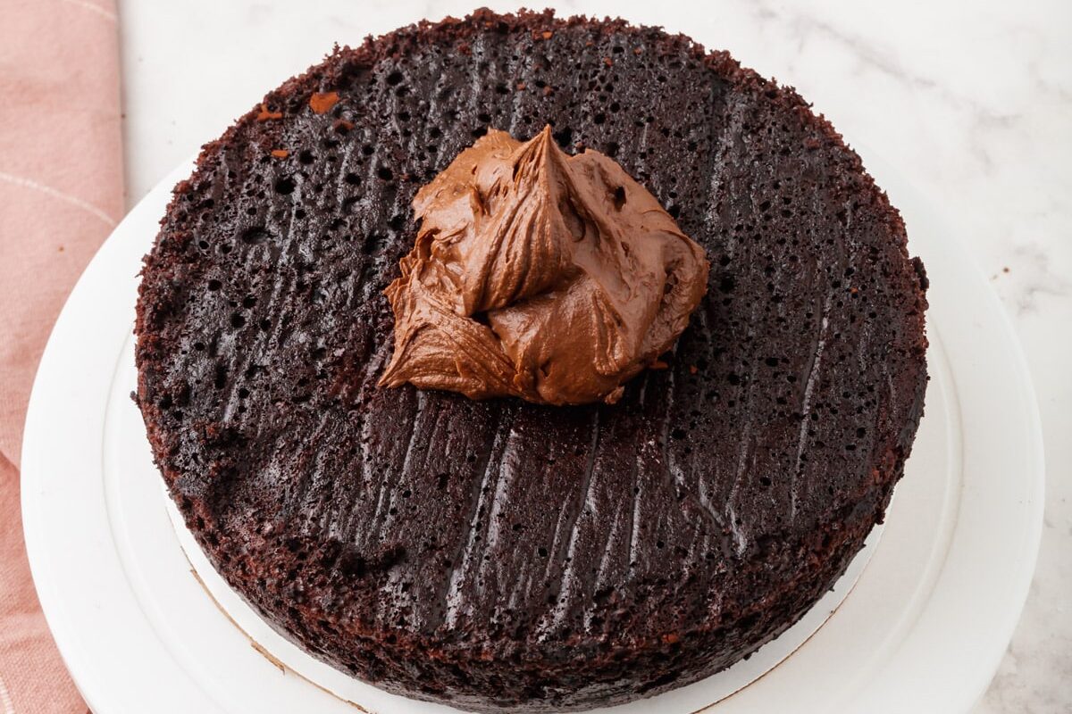 A single round chocolate cake layer sits on a white cake stand with a large dollop of chocolate frosting on top; a pink cloth is partially visible on the side, all on a marble surface.
