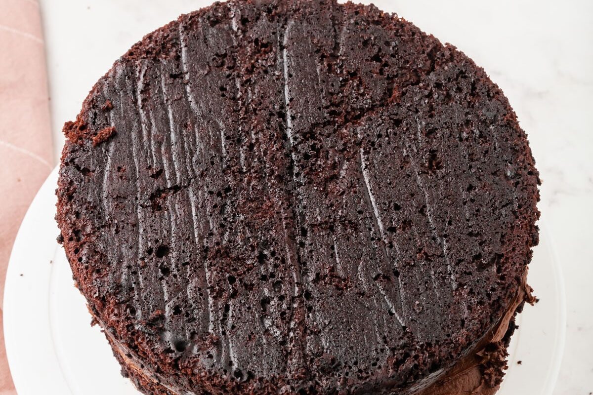 A round, unfrosted, two-layer chocolate cake with visible chocolate frosting between the layers, sits on a white cake stand atop a marble surface. A pink cloth is partially visible on the left.