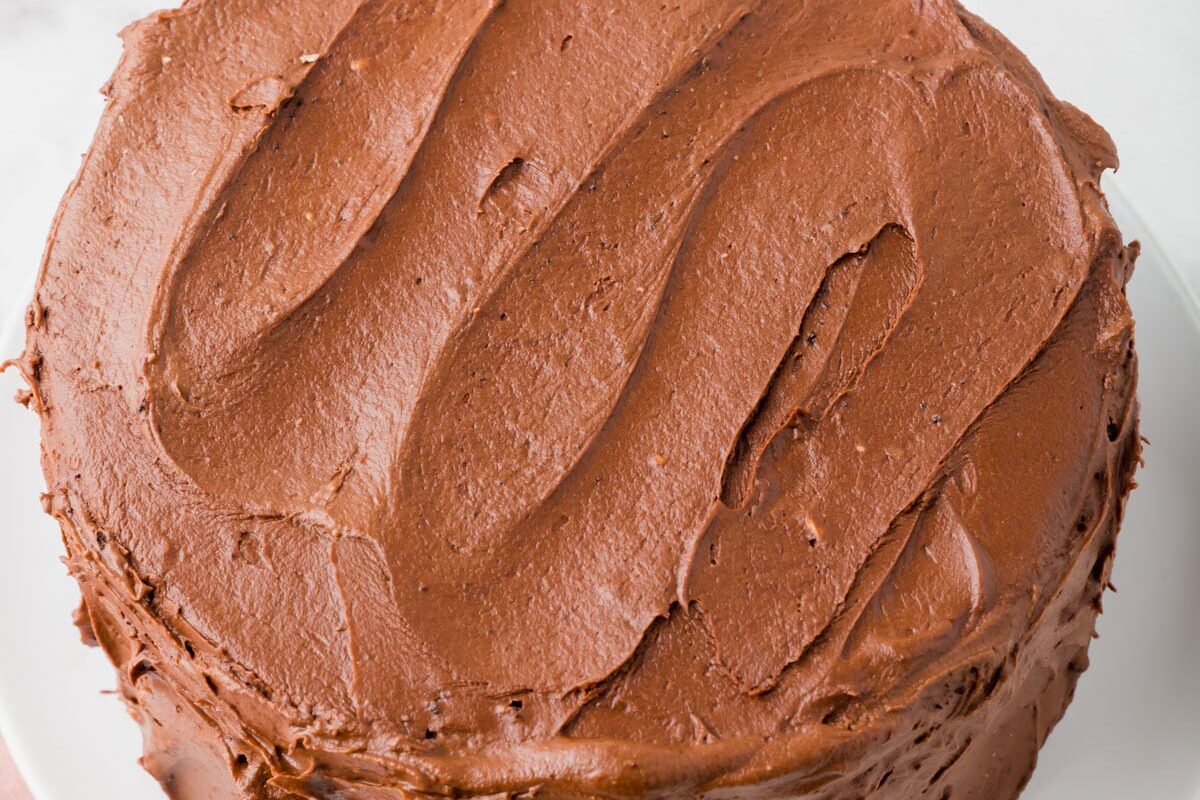 A round chocolate cake covered in smooth chocolate frosting sits on a white plate, with a pink cloth nearby and a cake server partially visible on the side.