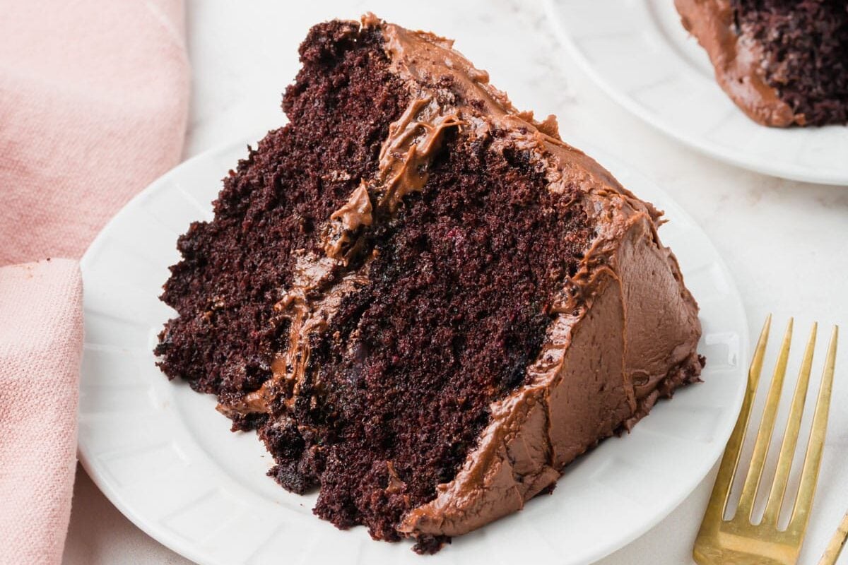 A slice of rich chocolate cake with chocolate frosting sits on a white plate beside a gold fork, with another slice and a pink napkin nearby on a marble surface.
